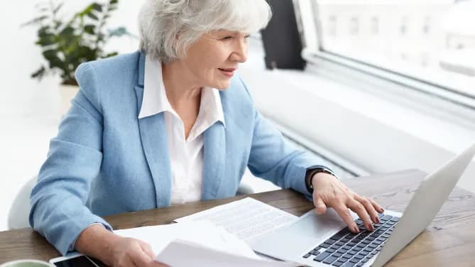 Woman typing on laptop with papers on desk in front of her and in her hand.