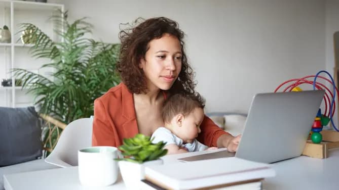 Woman sitting at a desk with a baby on her lap. There is a notebook and laptop in front of her on the table.