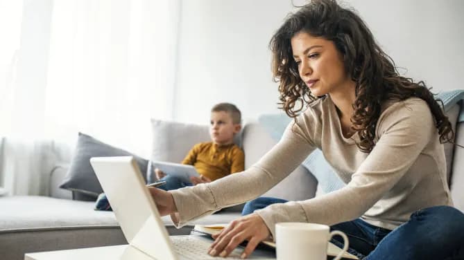 Woman on couch, working on laptop with a child sitting in the background.
