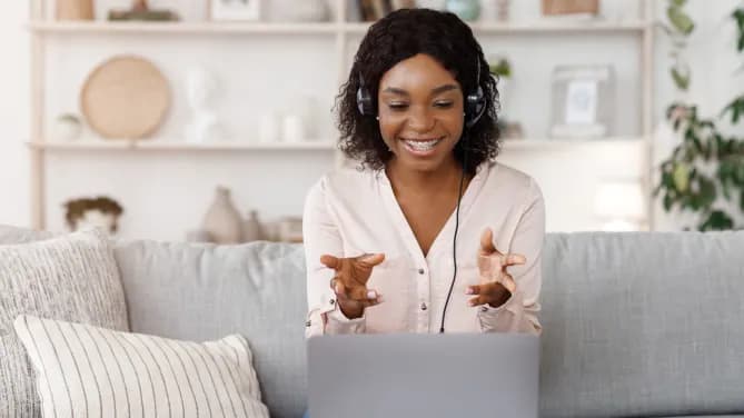 Woman with headphones on gray couch with laptop in front of her, smiling and talking with her hands.