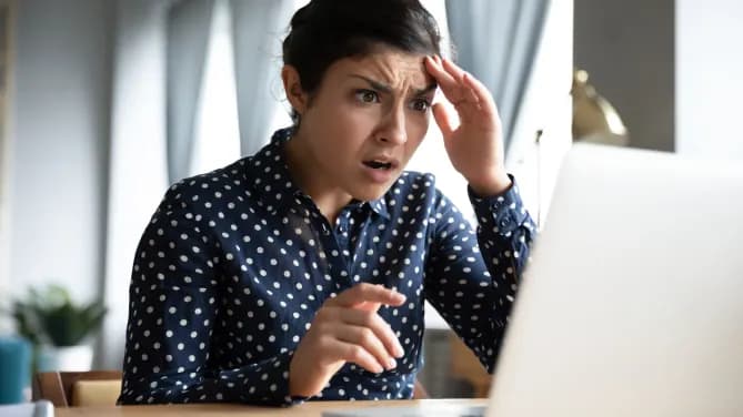Woman in blue polka dot shirt looks shocked at laptop.