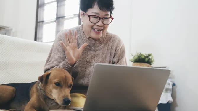 Woman sits on a couch next to her dog, waving at the computer in front of her.