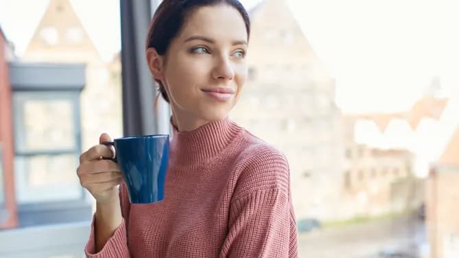 A woman smiles while holding a mug and leaning against a window